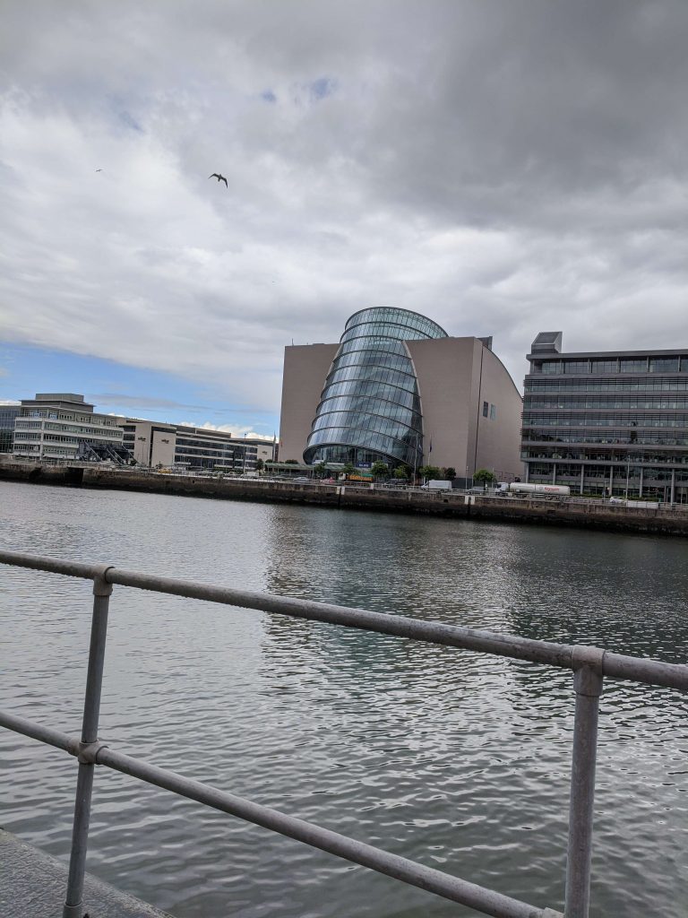 A photo taken from the opposite side of the canal of the Convention Centre Dublin. The building consists of a square block of concrete and a tilted cylinder of glass.
